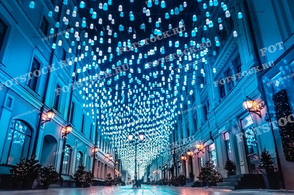 На фотографии изображена одна из центральных улиц Москвы, украшенная гирляндами в честь новогодних праздников. Синие и белые огни, натянутые над улицей, создают эффект сказочной атмосферы, освещая архитектуру зданий. На тротуарах видны небольшие ёлки, также украшенные в праздничной тематике. Ночная иллюминация отражает дух праздника и оживляет зимнюю столицу. Такая декорация улиц Москвы является неотъемлемой частью новогоднего убранства города.